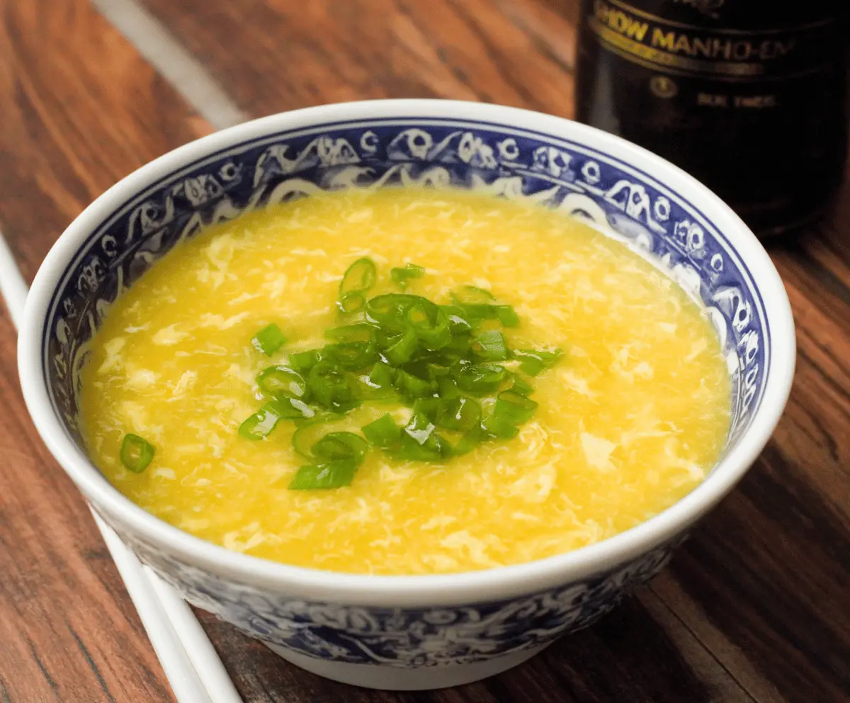 A steaming bowl of Egg Drop Soup garnished with chopped green onions and served in a white ceramic bowl, highlighting its savory broth and silky egg ribbons.